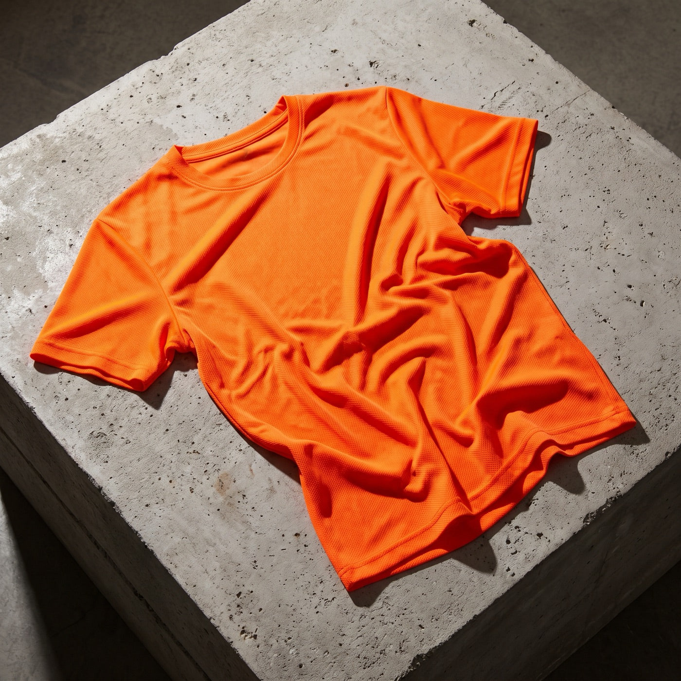 Bright orange athletic tee crumpled on raw concrete under harsh studio light